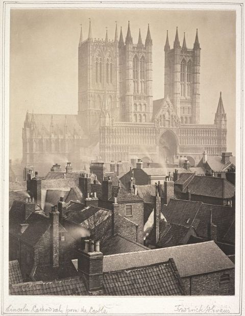Black and white photograph. In tones of warm brown and grey, the pointed towers and spires of a cathedral rise high above the nearby rooftops. Across the town from us, the church is faint, almost otherworldly, in the distance.