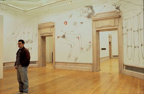 Installation photograph. An oversized line drawing of a nude man appears to have been created directly on the wall between two doors surrounded by tan-coloured moulding. He takes up the whole wall, his body, arms, and hands filling the space between and to either side of the doors. Touches of red scattered around his body suggest sores.